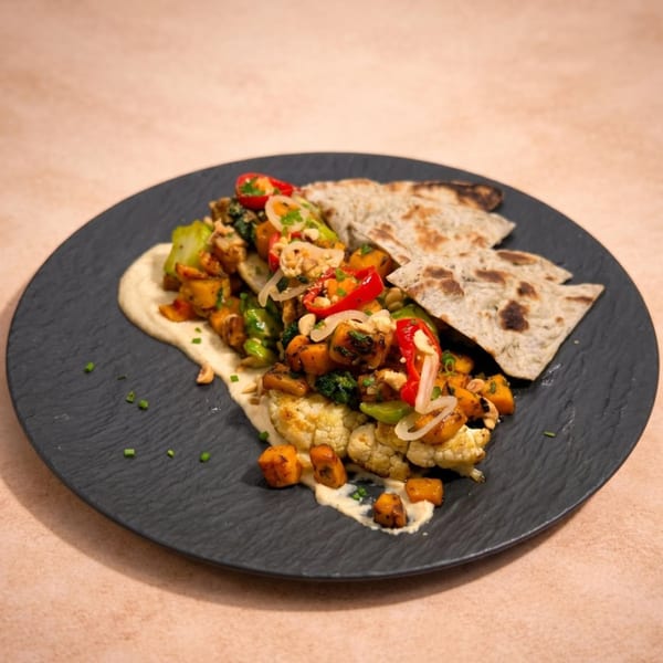 A black plate with roasted vegetables, hummus, and flatbread on a light brown surface.